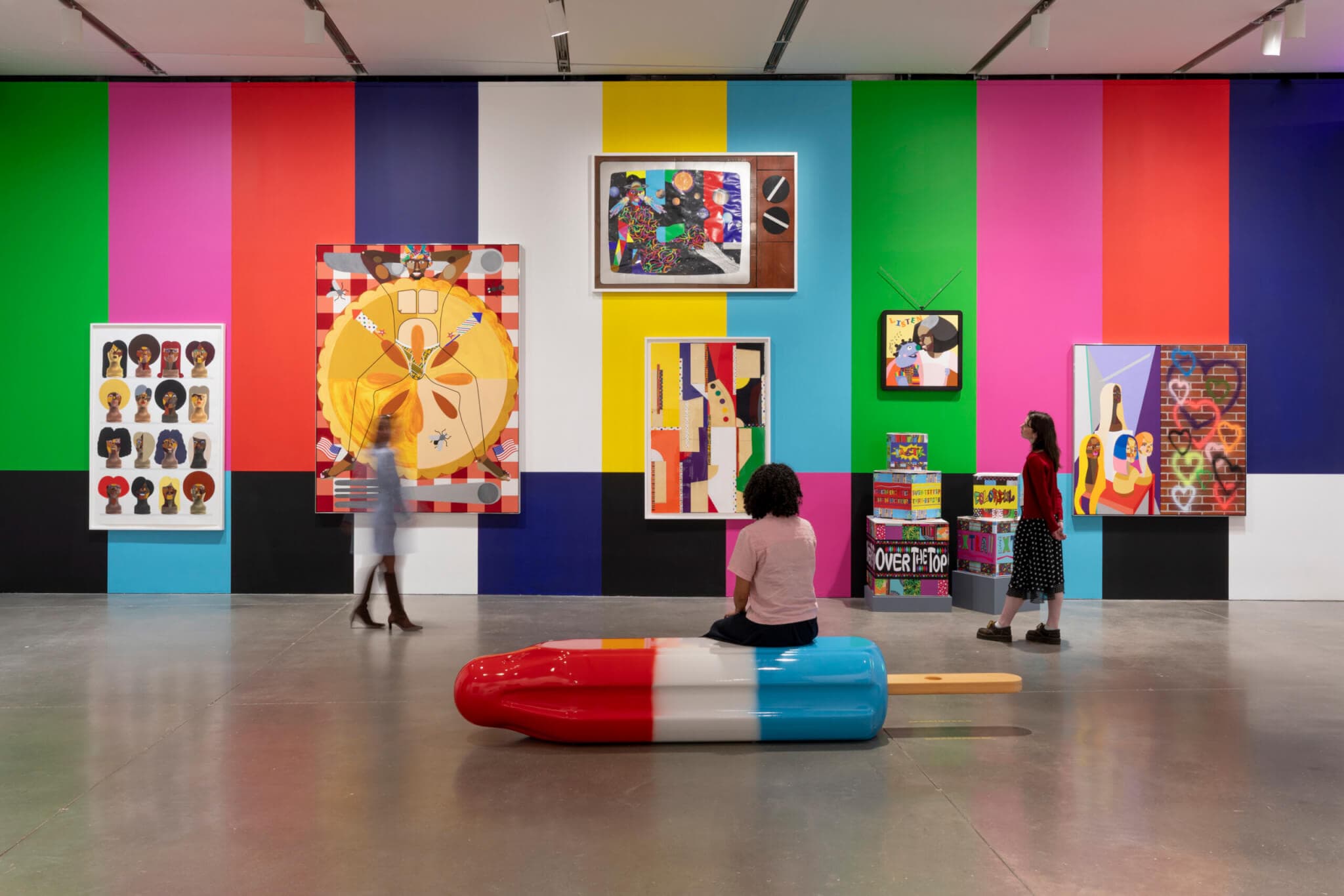 A woman sitting on a popsicle sculpture looks at framed artworks on a colorful striped wall.